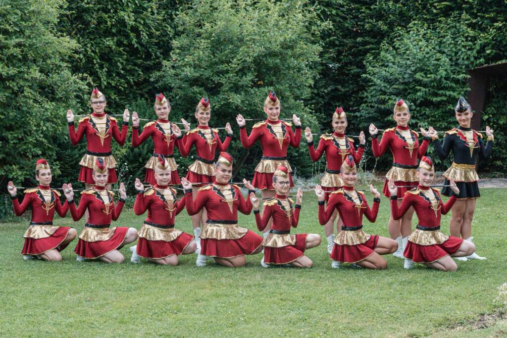 Juniorky v červeno-zlatých uniformách s čepičkami drží hůlky vodorovně nad rameny