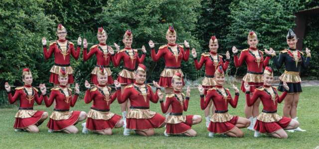 Juniorky v červeno-zlatých uniformách s čepičkami drží hůlky vodorovně nad rameny