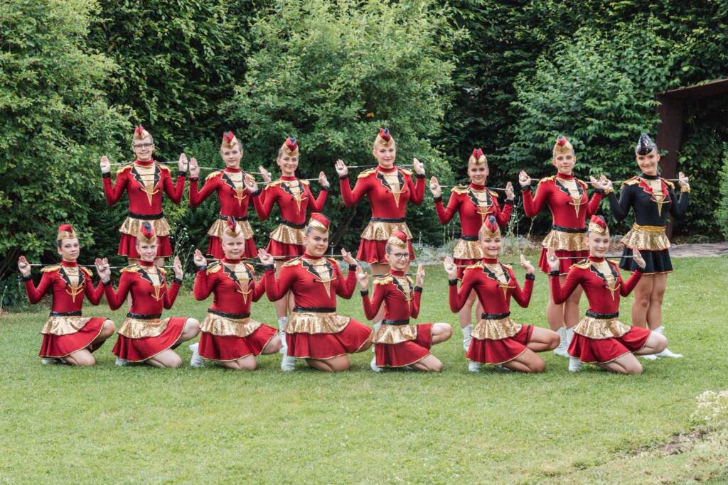 Červeno-zlatá formace juniorek SKP Nymburk ve dvou řadách, vedoucí v černém kostýmu